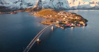 Karlı kayaların havadan görünüşü, deniz bölmesi, köprü, dağlar, yol, kışın gündoğumunda mavi gökyüzü. Norveç, Lofoten Adaları 'ndaki Hamnoy ve Reine köyünün güzel insansız hava aracı manzarası. Doğa