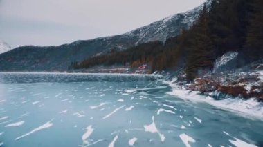 Kışın gün batımında karlı dağlarda donmuş gölün alçaktan uçan dronunun hava görüntüsü. Lofoten Adaları, Norveç. Buzlu nehrin üst manzarası, buzun üzerindeki yansıma, karlı kayalar, orman, karamsar gökyüzü. Doğa