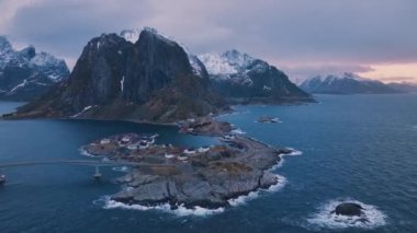 Karlı kayaların hava manzarası, deniz, köprü, dağ, yol, kışın gün doğumunda mor bulutlu gökyüzü. Norveç 'in Lofoten Adaları' ndaki Hamnoy köyünün insansız hava aracı görüntüsü. Dramatik