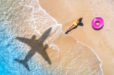 Uçak gölgesinin havadan görünüşü, yerde yatan güzel kadın, pembe yüzme halkası, tropik kumlu sahil, gün batımında dalgalı deniz. Adaya yaz tatili. Sıska kız manzarası, masmavi su, uçak gölgesi