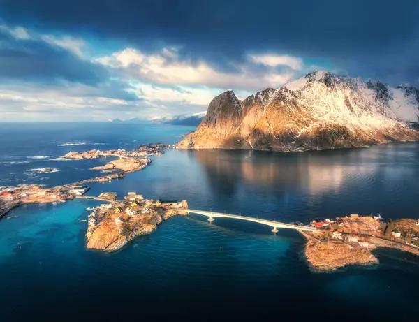 Aerial view of snowy rocks, island with rorbu, sea, bridge, water, mountains, road, cloudy sky at sunrise in winter. Beautiful dramatic landscape. Top view of Hamnoy village, Lofoten islands, Norway