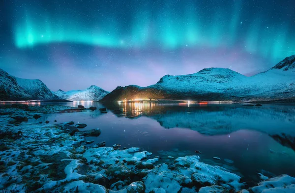 Aurora borealis over the snowy mountains, frozen sea, reflection in water at winter night in Lofoten, Norway. Northern lights and snowy rocks. Landscape with polar lights, stones, starry sky and fjord