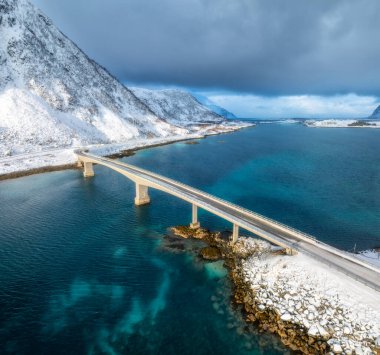 Norveç, Lofoten Adaları 'ndaki deniz üzerindeki köprülerin ve karlı dağların havadan görüntüsü. Soğuk kış gününde köprünün en iyi drone görüntüsü. Mavi suyu olan rüzgarlı bir manzara, kardaki kayalar, yol, bulutlu gökyüzü.