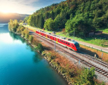 İlkbaharda gündoğumunda Alp Dağları 'nda nehir kıyısında hareket eden kırmızı, modern hızlı trenin havadan görünüşü. Tren, demiryolu, göl, yansıma, yazın yeşil ağaçlar. Slovenya 'daki tren istasyonu