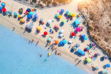 Kumlu sahildeki renkli şemsiyeler, güneşli yaz günlerinde mavi denizde yüzen insanlar. Sardunya, İtalya. Berrak gök mavisi suyu olan tropik bir yer. Seyahat ve tatil. Deniz kenarının üst görünümü