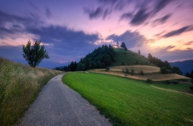 Gün batımında dağın zirvesinde kırsal yol ve şapel. Çakıl yolu, yeşil çayırlar ve ağaçlar tepedeki kilise ve Slovenya 'da alacakaranlıkta bulanık bulutlarla kaplı mor gökyüzü. Doğa. Yaz gecesi