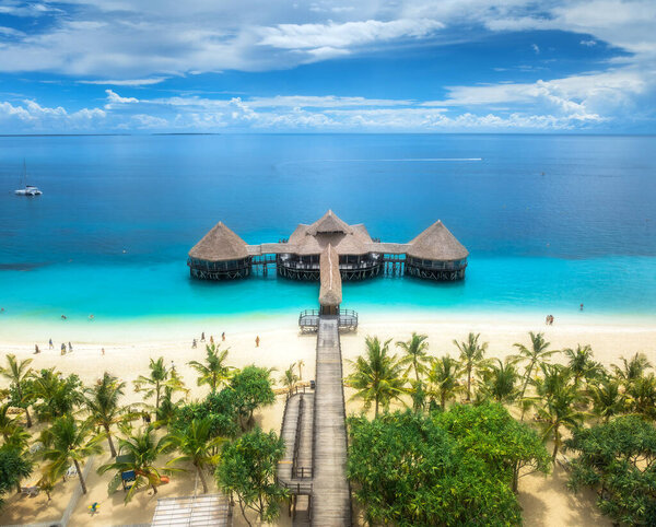 Aerial view of bungalow, white sandy beach, ocean on summer sunny day. Restaurant on the sea. Top drone view of wooden hotel, azure water, palms, sky with slouds. Luxury resort in Kendwa, Zanzibar