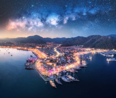 Parlak yıldızlı gökyüzünde parlayan Samanyolu Galaksisi 'nin ışıldayan tekneleriyle dolu canlı bir liman limanı olan bir kıyı şehrinin gece hava görüntüsü. Kasaba ışıklarının üst manzarası limanı aydınlatıyor. Marmaris, Türkiye