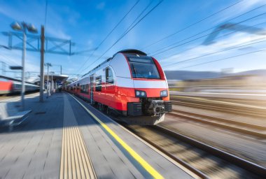 Yüksek hızlı kırmızı yolcu treni gün batımında açık mavi gökyüzünün altında tren istasyonunda hareket ediyor. Tren istasyonu. Bulanık hareket etkisine sahip modern demiryolu taşımacılığı konsepti. Demiryolu. Ticari.