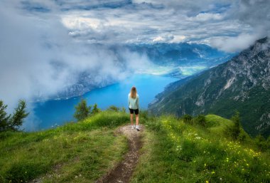 Dağın zirvesindeki patikada genç bir kadın ve yazın günbatımında alçak bulutlarda Garda Gölü 'nün güzel manzarası. Dolomitler, İtalya. Kız, patika, su, yeşil çimenler, sis ve gökyüzü ile renkli bir manzara. Yürüyüş