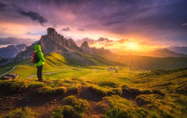 Sırt çantalı bir kadın ve yazın renkli günbatımında dağ zirveleri. Passo Giau, Dolomitler, İtalya. Kız manzarası, kayalar, tepeler, yeşil çimenler, çiçekler, güzel gökyüzü. Yürüyüş