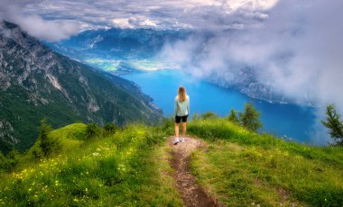 Dağın zirvesindeki patikada genç bir kadın ve yazın günbatımında alçak bulutlarda Garda Gölü 'nün güzel manzarası. Dolomitler, İtalya. Kız, patika, su, yeşil çimenler, sis ve gökyüzü ile renkli bir manzara. Yürüyüş