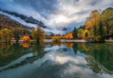Dağ Gölü, sonbahar günbatımında kırmızı ve turuncu yapraklı renkli ağaçları yansıtır. Suyla dramatik manzara, sisli orman, sonbaharda alçak bulutlarda dağlar. Jasna Gölü, Kranjska Gora, Slovenya