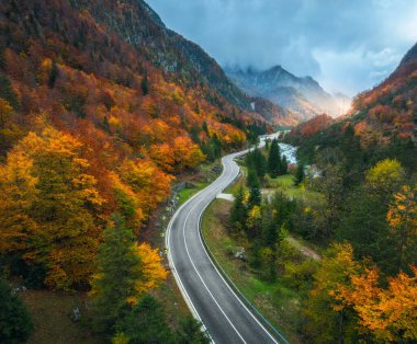 Gün batımında dağlardaki kıvrımlı yol ve turuncu ormanın havadan görünüşü. İtalya 'da altın sonbahar. Virajlı yolun üst manzarası, renkli ağaçlar, nehir, sonbaharda dağlar. Boş yol ve orman. Huysuz manzara