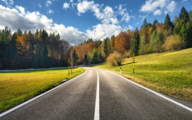 Yol, yeşil çayırlar ve turuncu orman altın sonbaharda gün batımında. İtalya. Güzel dağ yolu, ağaçlar, çimenler, bulutlu mavi gökyüzü. Sonbaharda otobanı, tarlası ve ormanı boş bir manzara. Seyahat