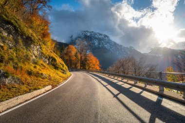 Sonbaharda gün batımında dağ yolu. Alpler, İtalya. Güzel kıvrımlı dağ yolu, karlı kayalar, ağaçlar, portakal yaprakları, bulutlu dramatik gökyüzü. Sonbaharda dağ geçidinde boş yolu olan renkli bir manzara.