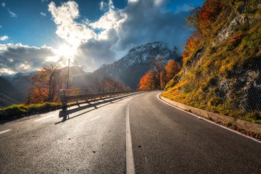 Sonbaharda gün batımında dolambaçlı dağ yolu. Alpler, İtalya. Güzel kıvrımlı dağ yolu, karlı kayalar, ağaçlar, kırmızı yapraklar, bulutlu mavi gökyüzü. Sonbaharda dağ geçidinde boş yolu olan renkli bir manzara.