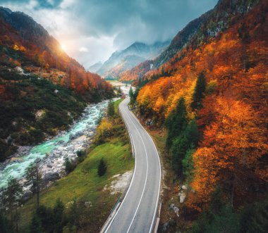 Gün batımında dağlardaki kıvrımlı yol ve turuncu ormanın havadan görünüşü. İtalya 'da altın sonbahar. Virajlı yolun üst manzarası, renkli kırmızı ağaçlar, nehir, sonbaharda dağlar. Boş yol, orman. Huysuz manzara