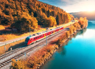 Sonbaharda gündoğumunda Alp Dağları 'nda göl kenarında hareket eden modern kırmızı hızlı trenin havadan görünüşü. Tren, demiryolu, su, yansıma, sonbaharda portakal ağaçları. Slovenya 'daki tren istasyonu