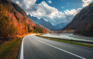 Sonbaharda gün batımında dağ yolu. Alpler, İtalya. Güzel kıvrımlı dağ yolu, karlı kayalar, ağaçlar, kırmızı yapraklar, nehir, bulutlu gökyüzü. Sonbaharda dağ geçidinde boş yolu olan renkli bir manzara.