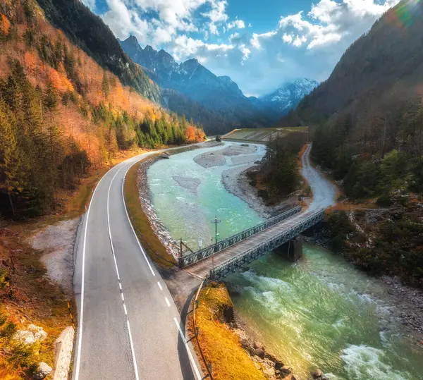 Sonbaharda günbatımında dönen dağ yolunun hava manzarası. Alpler, İtalya. Alp yolu, köprü, nehir, karlı kayalar, renkli ağaçlar, kırmızı yapraklar, göl, bulutlu gökyüzü. Sonbaharda dağın tepesindeki yol manzarası