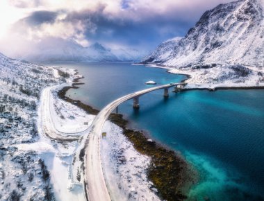 Norveç, Lofoten Adaları 'ndaki deniz üzerindeki köprülerin ve karlı dağların havadan görüntüsü. Kışın gün batımında köprü manzarası. Mavi suyu olan rüzgarlı bir manzara, kardaki kayalar, yol, bulutlu gökyüzü.