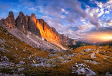 Sonbaharda güzel günbatımında dağlar. Passo Sella, Dolomitler, İtalya. Dağ zirveleri, kayalar, alp çayırları, taşlar, patikalar, turuncu çimenler ve çiçekler, sonbaharda bulutlarla kaplı gökyüzü.