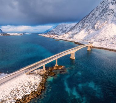 Norveç, Lofoten Adaları 'ndaki deniz üzerindeki köprülerin ve karlı dağların havadan görüntüsü. Kışın gün batımında köprü manzarası. Mavi suyu olan rüzgarlı bir manzara, kardaki kayalar, yol, bulutlu gökyüzü.