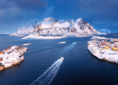 Norveç 'in Lofoten Adaları' ndaki Henningsvaer köyündeki mavi denizde yüzen balıkçı teknesinin hava manzarası. Geminin üst drone görüntüsü, karlı dağlar, deniz, bulutlu gökyüzü, kasaba, hata kesici ve evler