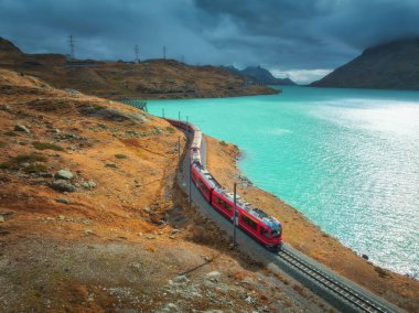Gök mavisi göl kenarındaki modern kırmızı trenin havadan görünüşü ve sonbaharda günbatımında dramatik bulutlu gökyüzü. Bernina Express, İsviçre. Alplerdeki tren manzarası, dolambaçlı demiryolu, sonbaharda turuncu tepeler.