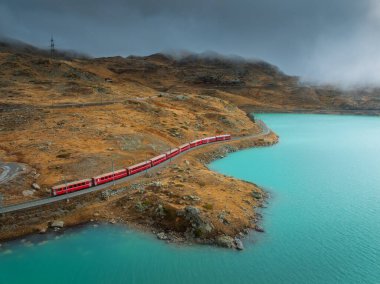 Gök mavisi göl kenarındaki modern kırmızı trenin havadan görünüşü ve sonbaharda günbatımında dramatik bulutlu gökyüzü. Bernina Express, İsviçre. Alplerdeki tren manzarası, dolambaçlı demiryolu, sonbaharda turuncu tepeler.