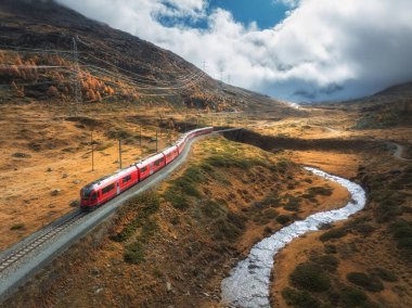 Modern kırmızı trenin, nehrin, dağların, turuncu tepelerin ve ağaçların, sonbaharda günbatımında bulutlu gökyüzünün havadan görünüşü. Bernina Express, İsviçre. Alplerdeki trenin en üst manzarası, sonbaharda demiryolu dolambaçlı.