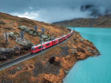 Gök mavisi göl kenarındaki modern kırmızı trenin havadan görünüşü ve sonbaharda günbatımında dramatik bulutlu gökyüzü. Bernina Express, İsviçre. Alplerdeki tren manzarası, dolambaçlı demiryolu, sonbaharda turuncu tepeler.