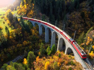 Landwasser 'daki modern kırmızı trenin sonbaharda günbatımında turuncu orman ve dağlardaki viyadük manzarası. Bernina Express, İsviçre. Alplerdeki tren manzarası, demiryolu, sonbaharda renkli ağaçlar.
