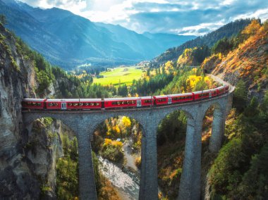 Landwasser 'daki modern kırmızı trenin sonbaharda günbatımında turuncu orman ve dağlardaki viyadük manzarası. Bernina Express, İsviçre. Alplerdeki tren manzarası, demiryolu, sonbaharda renkli ağaçlar.