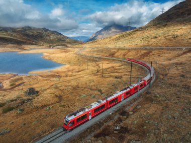 Sonbaharda günbatımında bulutlarla birlikte dağ gölü ve gökyüzü yakınlarındaki modern kırmızı trenin hava manzarası. Bernina Express, İsviçre. Alplerdeki hız treni manzarası, dolambaçlı demiryolu, sonbaharda turuncu tepeler..