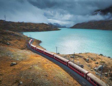 Gök mavisi göl kenarındaki hareket eden kırmızı trenin ve sonbaharda günbatımında dramatik bulutlu gökyüzünün görüntüsü. Bernina Express, İsviçre. Alplerdeki tren manzarası, dolambaçlı demiryolu, sonbaharda turuncu tepeler.
