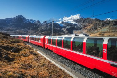 Sonbaharda mavi gökyüzü olan güneşli bir günde İsviçre Alp Dağları 'nda kırmızı tren demiryolu boyunca ilerliyor. Bernina Ekspres treni, turuncu çimenler, sonbaharda gün batımında karlı kayalar. Seyahat