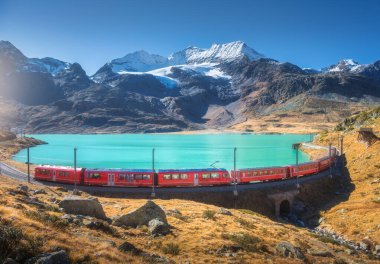 Gök mavisi Bianco Gölü yakınlarındaki modern kırmızı trenin havadan görünüşü, sonbaharda günbatımında karlı kayalar ve mavi gökyüzü. Bernina Express, İsviçre. Alplerdeki tren manzarası, demiryolu, sonbaharda turuncu tepeler