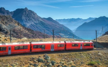 Kızıl modern tren güneşli bir sonbahar gününde İsviçre Alpleri 'nden geçiyor. Hareket eden tren, dağlık vadi, turuncu tepeler, sonbaharda gün batımında mavi gökyüzü. Bernina Ekspresi, İsviçre