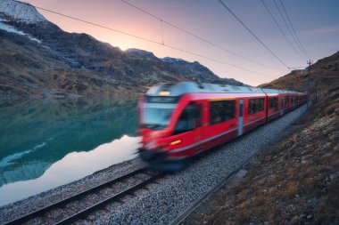 Gök mavisi dağ Bianco Gölü 'nün yanındaki modern kırmızı tren, karlı kayalar ve sonbaharda günbatımında renkli gökyüzü. Bernina Express, İsviçre. Alplerde bulanık tren, alacakaranlıkta suda yansıma.