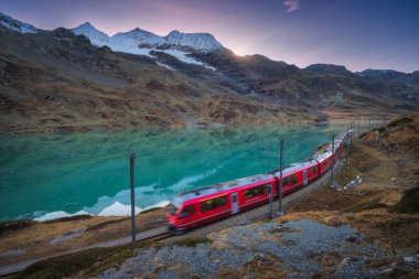 Gök mavisi dağ Bianco Gölü 'nün yanındaki modern kırmızı tren, karlı kayalar ve sonbaharda günbatımında renkli gökyüzü. Bernina Express, İsviçre. Alplerde bulanık tren, alacakaranlıkta suda yansıma.