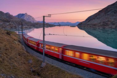 Gök mavisi dağ Bianco gölü yakınlarındaki modern kırmızı tren, karlı kayalar ve sonbaharda günbatımında renkli pembe gökyüzü. Bernina Express, İsviçre. Alplerde bulanık tren, alacakaranlıkta suda yansıma.