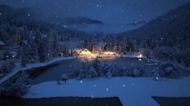 Kar yağışı sırasındaki peri ormanı manzarası, donmuş göl ve kışın geceleri ışıkları olan ev. Dağ köyü, gölet, aydınlatma, karlı çam ağaçları, Slovenya 'da alacakaranlıkta bulutlar.