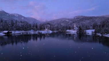 Kışın kar yağarken göl manzarası. Karlı dağlar, ağaçlar, kilise, köprü, yol, şehir ışıkları, evler, sudaki yansıma, mor gökyüzü. Alacakaranlık. Bohinj, Slovenya