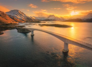 Kışın günbatımında Fredvang köprülerinin manzaralı manzarası. Norveç, Lofoten takımadalarındaki adalar. Köprü manzarası, karla kaplı dağlar, deniz, turuncu gökyüzü, suda yansıma.