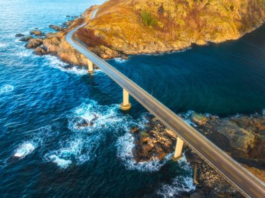 Moskenes adasını anakaraya bağlayan djupfjordbrua köprüsünün havadan görünüşü, Lofoten takımadasındaki mavi okyanus suyu ve kayalık kıyı şeridi, Norveç, gün batımında. Yol manzarası, denizdeki kayalar