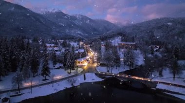 Kışın kar yağarken göl manzarası. Karlı dağların, ağaçların, köprünün, arabaların, yolun, şehrin ışıklarının, evlerin, sudaki yansımanın, alacakaranlıkta mor gökyüzünün en üst drone görüntüsü. Bohinj, Slovenya