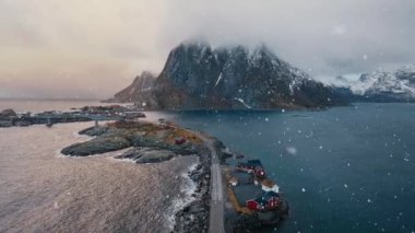 Karlı dağların havadan görünüşü, rorbu evlerinin olduğu adalar, mavi deniz, köprü, yol, kış günbatımında bulutlu gökyüzü. Norveç 'in Lofoten Adaları' ndaki Hamnoy köyünün insansız hava aracı görüntüsü. Dramatik