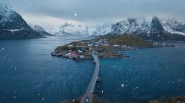 Karlı dağların havadan görünüşü, daha büyük evleri olan adalar, mavi deniz, köprü, yol, kışın bulutlu gökyüzü. Norveç 'in Lofoten Adaları' ndaki Hamnoy köyünün insansız hava aracı görüntüsü. Dramatik